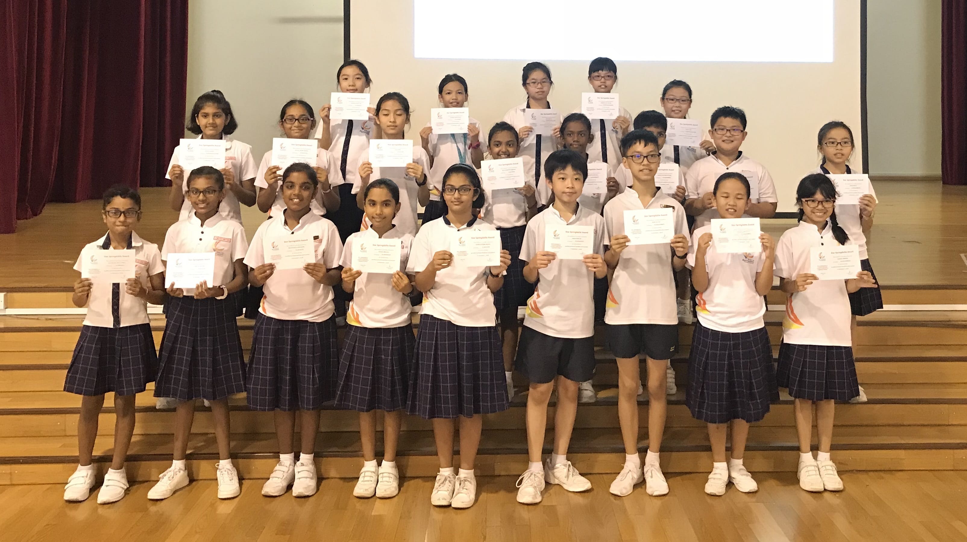 A group of school children in white uniforms standing on stage steps, each holding a certificate.